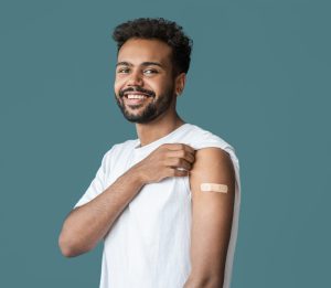 Young man pulling up sleeve in white shirt to show bandaid after receiving a vaccine, with teal background
