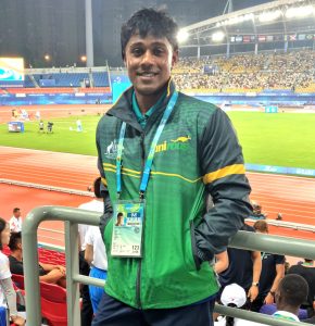 Dr Sharujan Sivasubramaniam (SJ) Sports Medicine Doctor, in green, navy and yellow uniroos jersey leaning against railing, smiling with hands in pockets at stadium